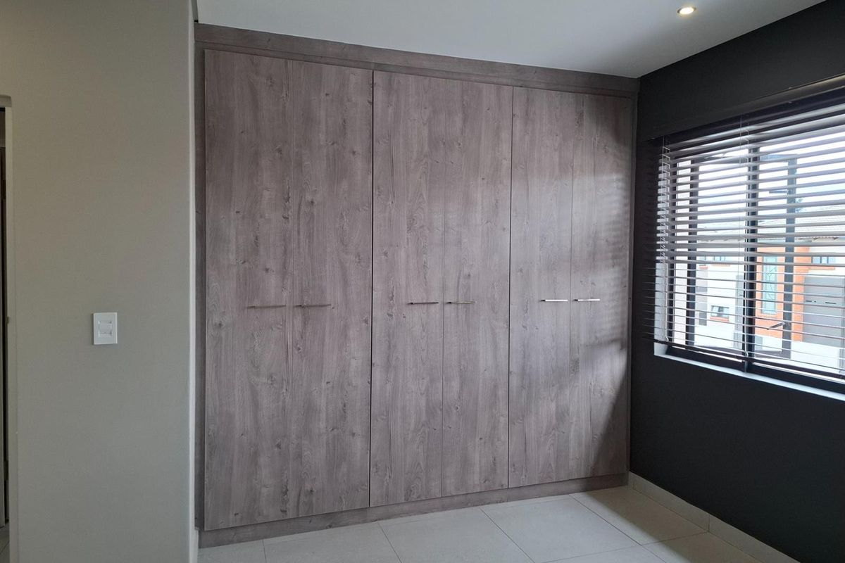A modern bedroom featuring floor-to-ceiling built-in wooden cupboards, tiled flooring, and a window fitted with horizontal blinds.