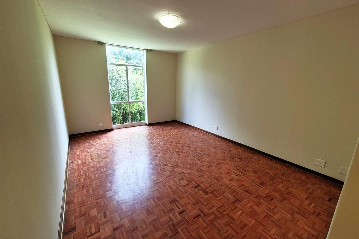 A bright, empty bedroom featuring classic parquet wood flooring, neutral walls, and a large window providing natural light and a green outlook.