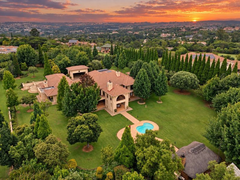 Aerial view of a large residential property featuring a spacious landscaped garden, swimming pool, covered patio, and extensive mature trees at sunset.