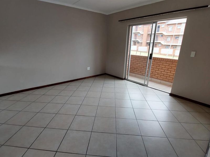 An empty open-plan living area featuring tiled flooring and a sliding glass door leading to a balcony, with an adjacent kitchen corner.
