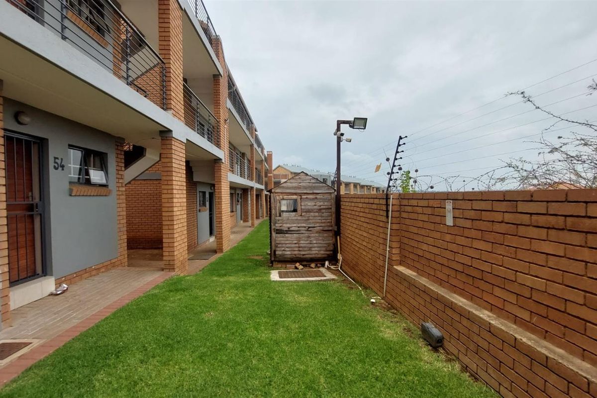 Rear exterior view of the townhouse complex featuring a private lawn area, brick boundary wall with electric fencing, and access to upper-level balconies.