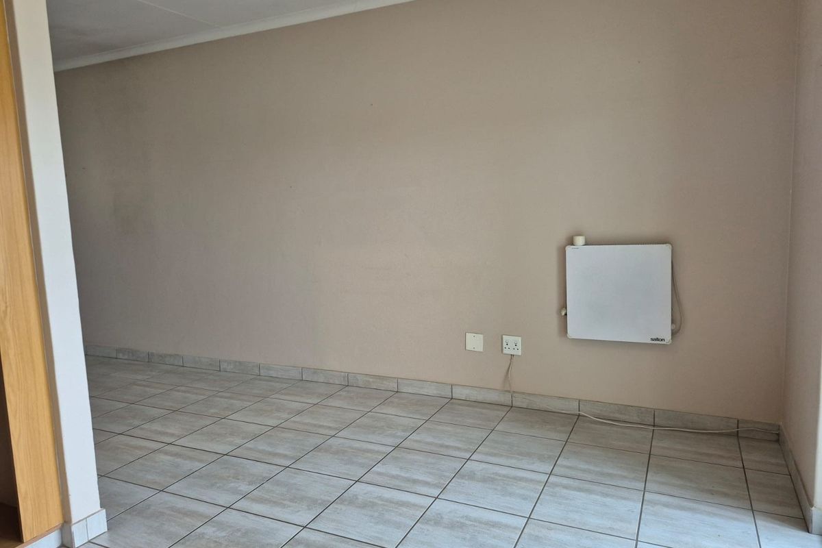 An empty living area featuring neutral tiled flooring, a wall-mounted heater, and standard electrical outlets.