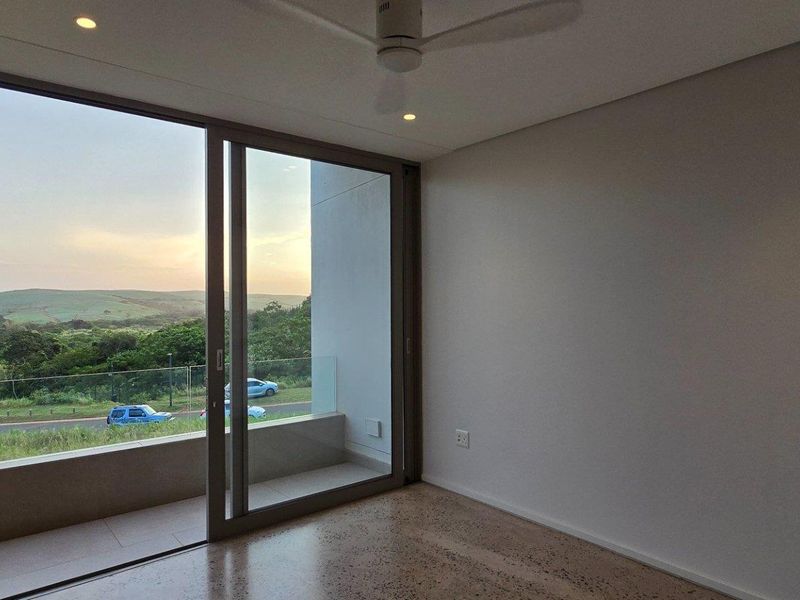 A modern, bright bedroom featuring polished concrete flooring, a ceiling fan, and large sliding glass doors opening onto a private balcony with scenic views.