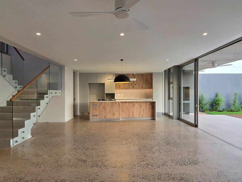 Spacious open-plan living area featuring a modern kitchen with wood-finish cabinetry, polished concrete flooring, and a glass-railed staircase leading to the upper level.