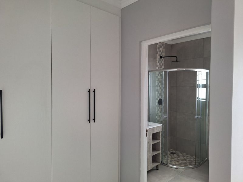 A view from a bedroom featuring built-in cupboards leading into a modern en-suite bathroom with a glass-enclosed shower and contemporary tiling.