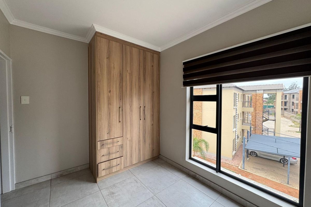 A bright bedroom featuring modern wood-finish built-in cupboards, large windows with fitted blinds, and neutral floor tiling.