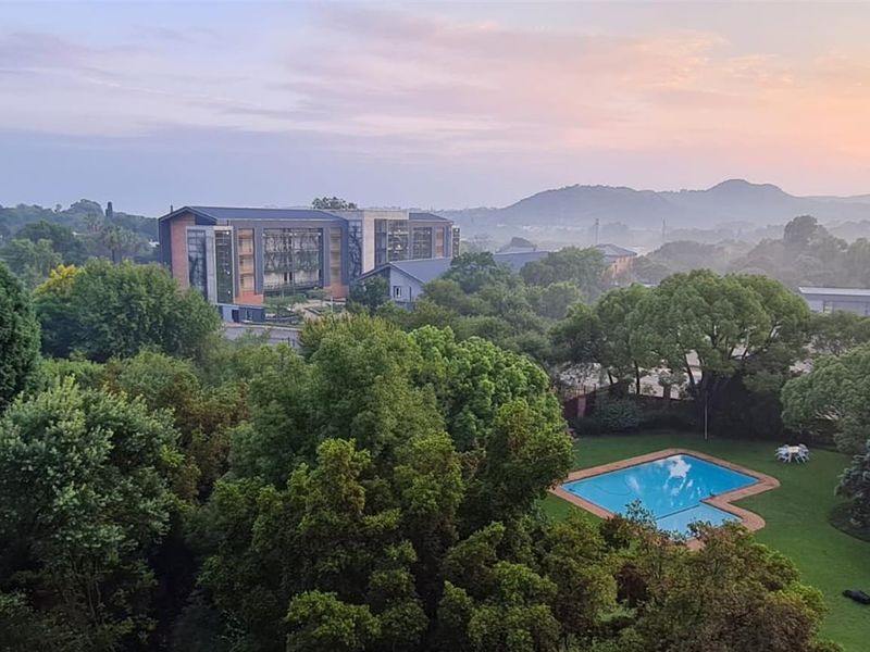 Elevated scenic view overlooking a lush green landscape, mature trees, and a communal swimming pool area at sunset.