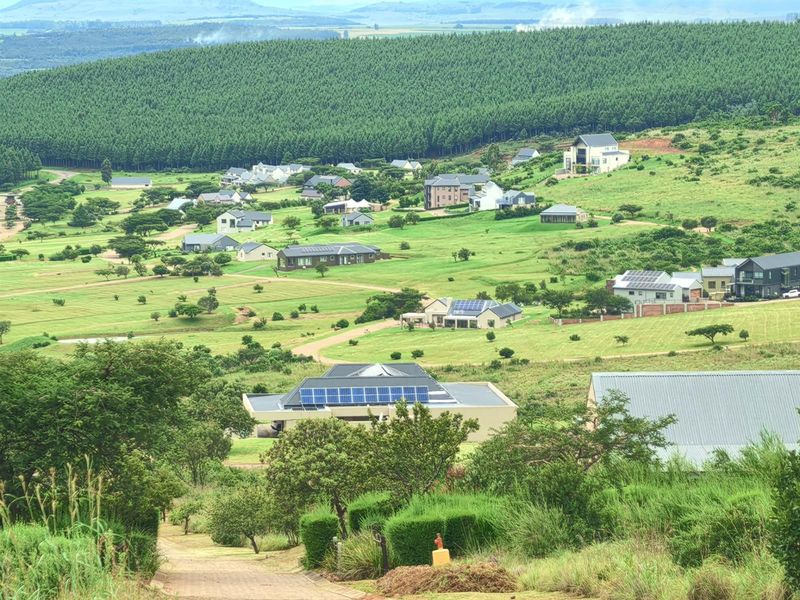 An elevated aerial view of a residential estate featuring multiple homes, paved roads, and expansive green landscapes with surrounding forest.