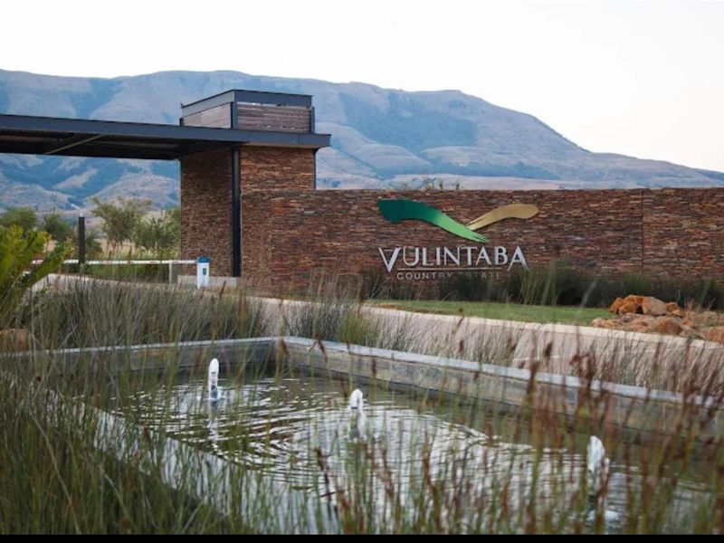 The entrance to Vulintaba Country Estate featuring a stone-clad wall, water feature, and security gatehouse set against a scenic mountain backdrop.