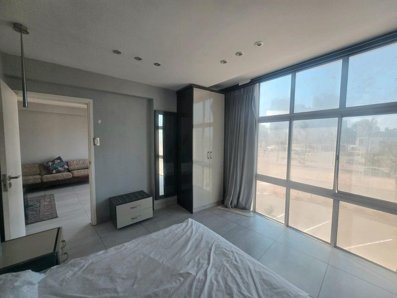 A bright bedroom featuring large floor-to-ceiling windows, built-in wardrobe storage, recessed ceiling lighting, and neutral tiled flooring.