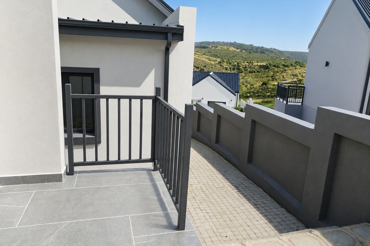 A modern exterior rear view featuring a tiled patio with a metal balustrade, a paved walkway, and a clean boundary wall.