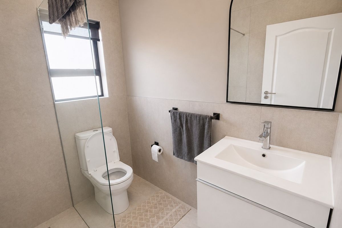 Modern bathroom featuring a sleek vanity, wall-mounted mirror, toilet, and a glass-enclosed shower area with neutral floor-to-ceiling tiling.