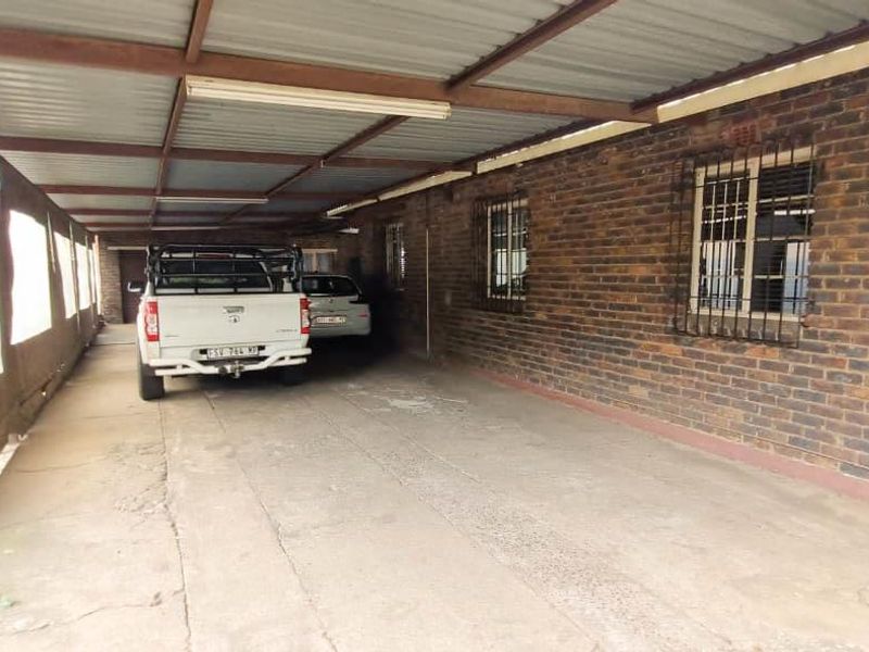 A covered carport area featuring exposed brick walls, security-barred windows, and a corrugated metal roof structure with a concrete driveway.