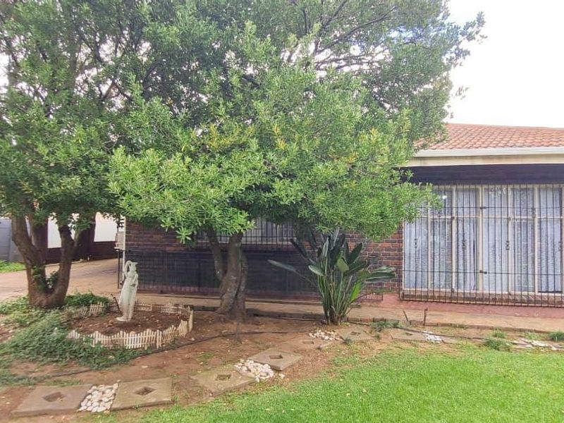 Front elevation of a brick house featuring a tiled roof, security-barred sliding doors, and a landscaped garden area with mature trees.
