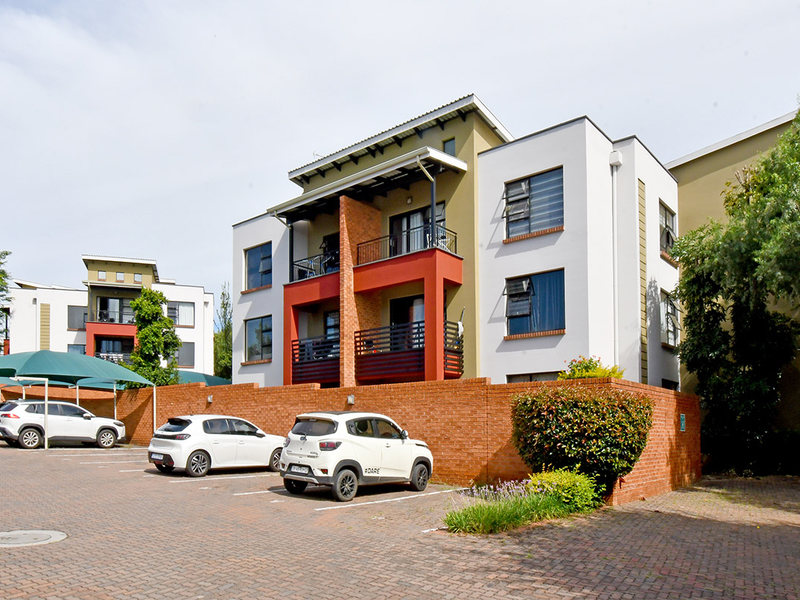 Exterior view of a modern apartment complex featuring brick boundary walls, paved parking with shade cloth carports, and private balconies with metal railings.