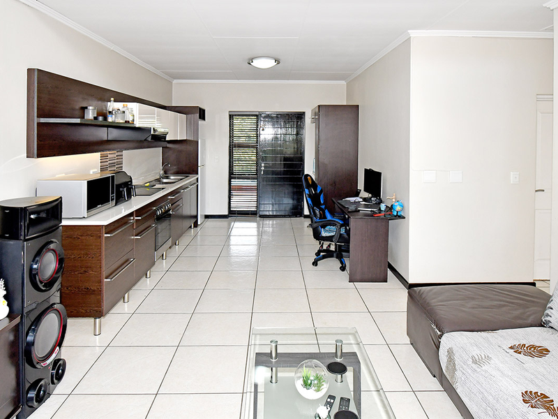 Open-plan living area featuring tiled flooring and a functional kitchen with built-in cabinetry, oven, and extractor fan, leading to an exterior security door.