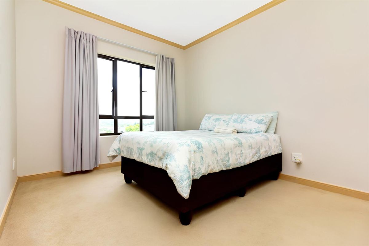 A well-lit bedroom featuring neutral carpeting, beige walls with crown molding, and a large window providing natural light.