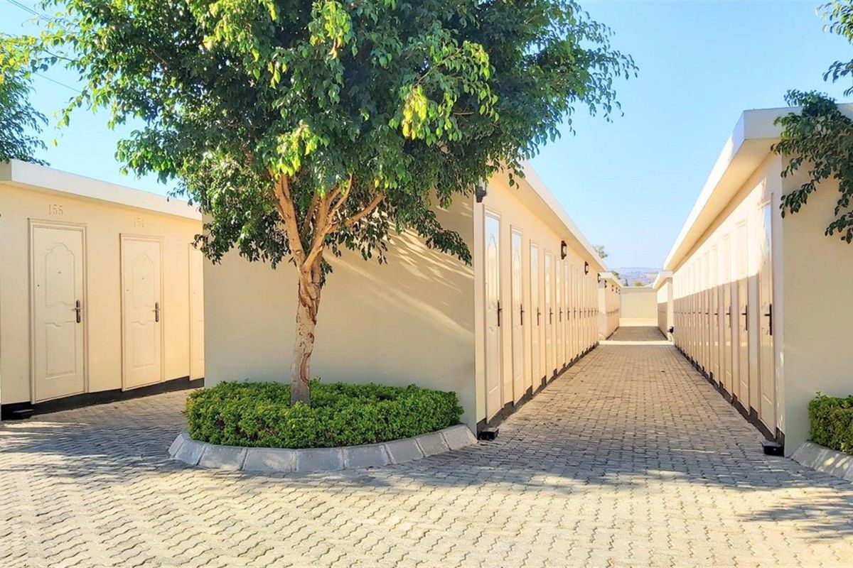 Exterior view of a residential apartment complex featuring paved walkways, uniform unit doors, and landscaped garden beds with mature trees.