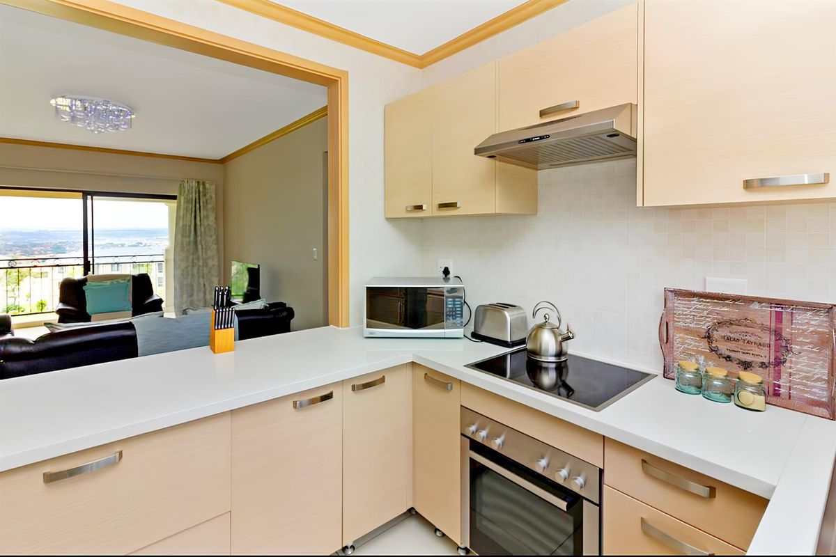 A compact, modern kitchen featuring light wood cabinetry, a built-in oven, electric hob, and extractor fan, opening into a living area with balcony access.