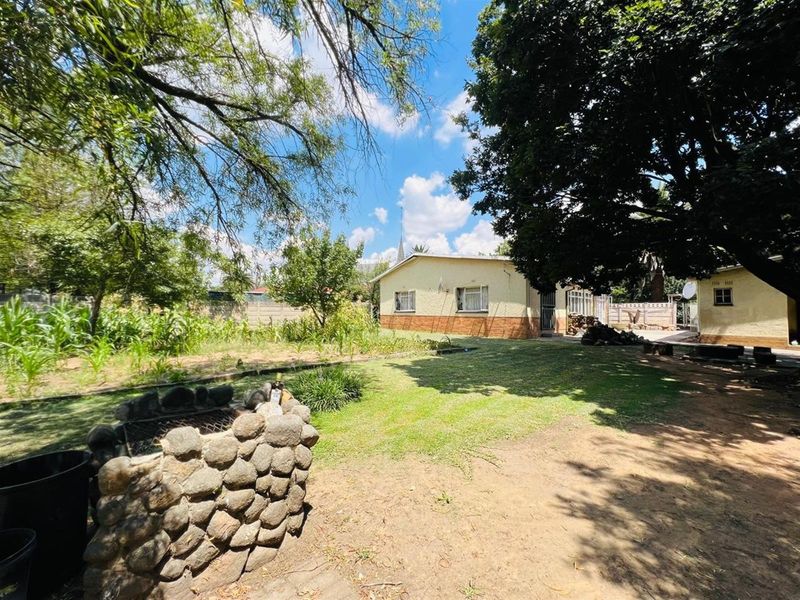 A spacious rear garden featuring a stone-built braai area, mature trees, and a view of the property's rear elevation.