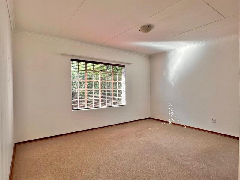 A spacious, empty bedroom featuring neutral carpeting, a large window with security bars, and a simple ceiling-mounted light fixture.