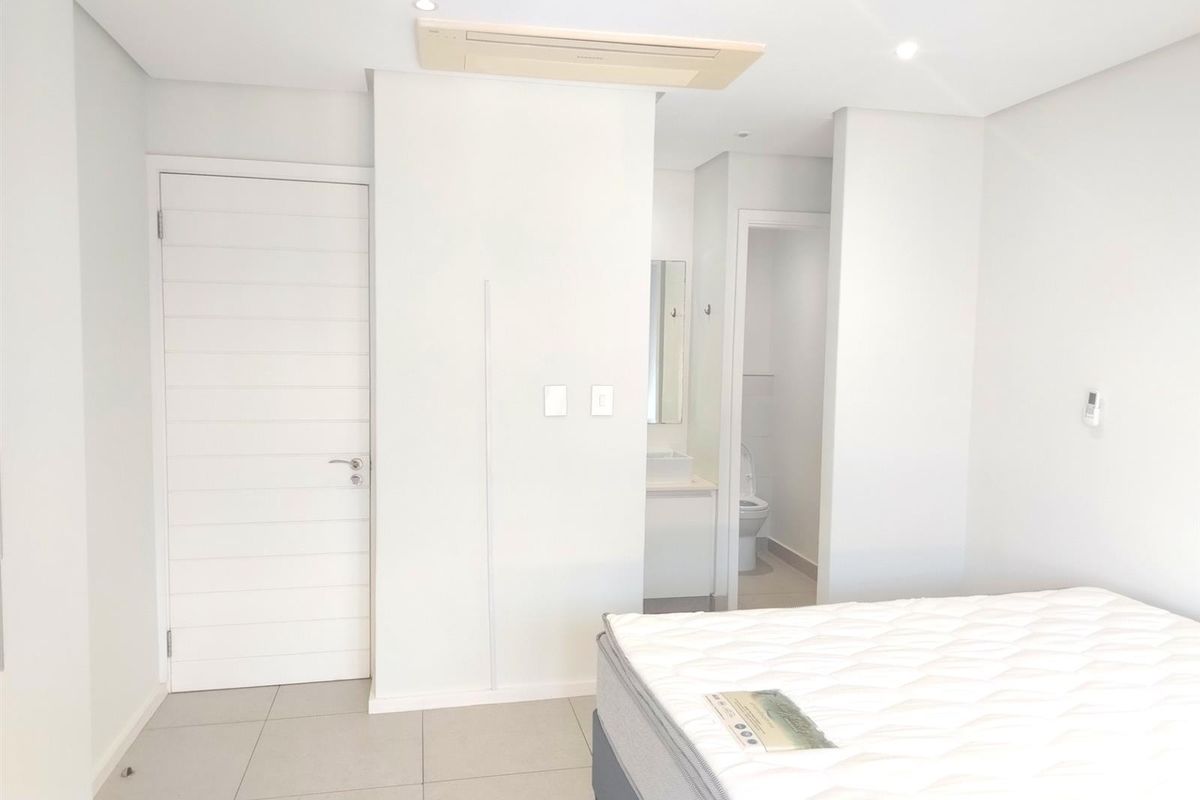A bright bedroom featuring neutral tiled flooring, recessed ceiling lighting, a built-in air conditioning unit, and an en-suite bathroom.