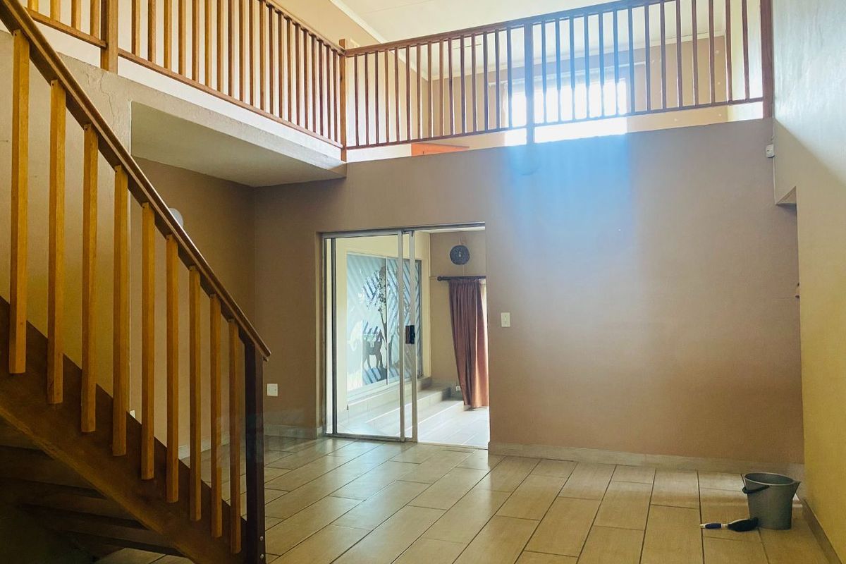 Spacious double-volume living area featuring a wooden staircase, mezzanine landing with timber balustrades, tiled flooring, and sliding glass doors leading to an outdoor space.