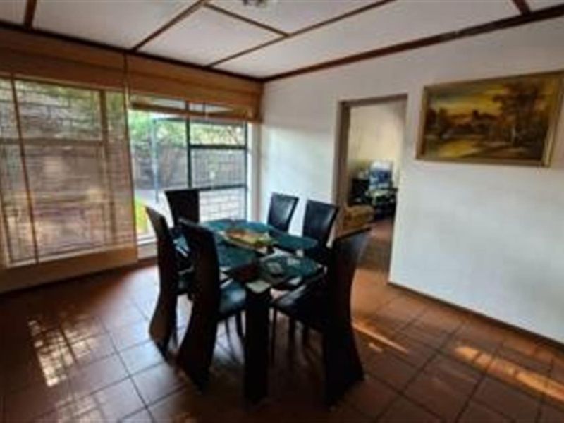 A dining room featuring tiled flooring and an exposed beam ceiling, with large windows providing natural light and a view of the exterior.