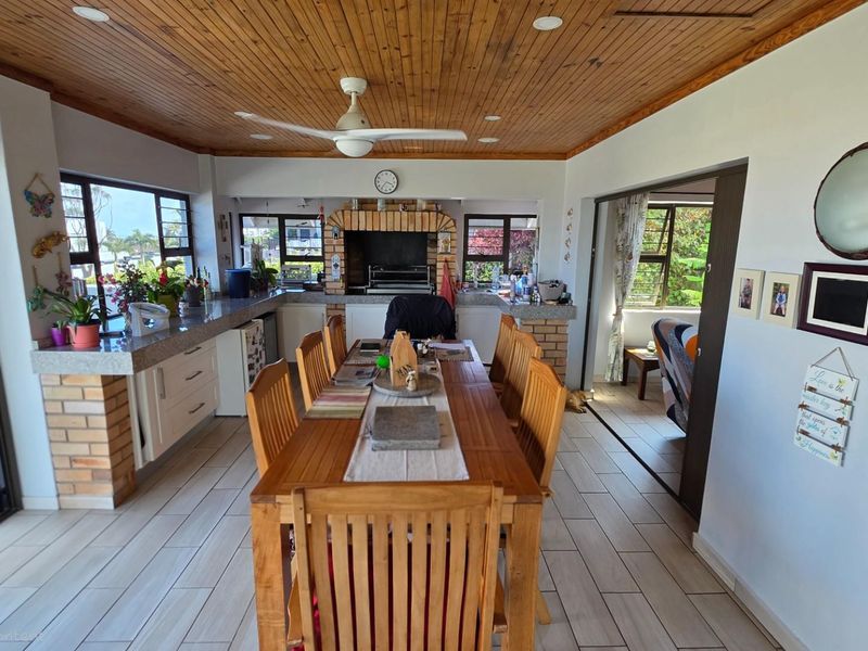 Spacious open-plan entertainment area featuring a built-in brick braai, granite countertops, timber-clad ceiling, and large windows providing natural light.