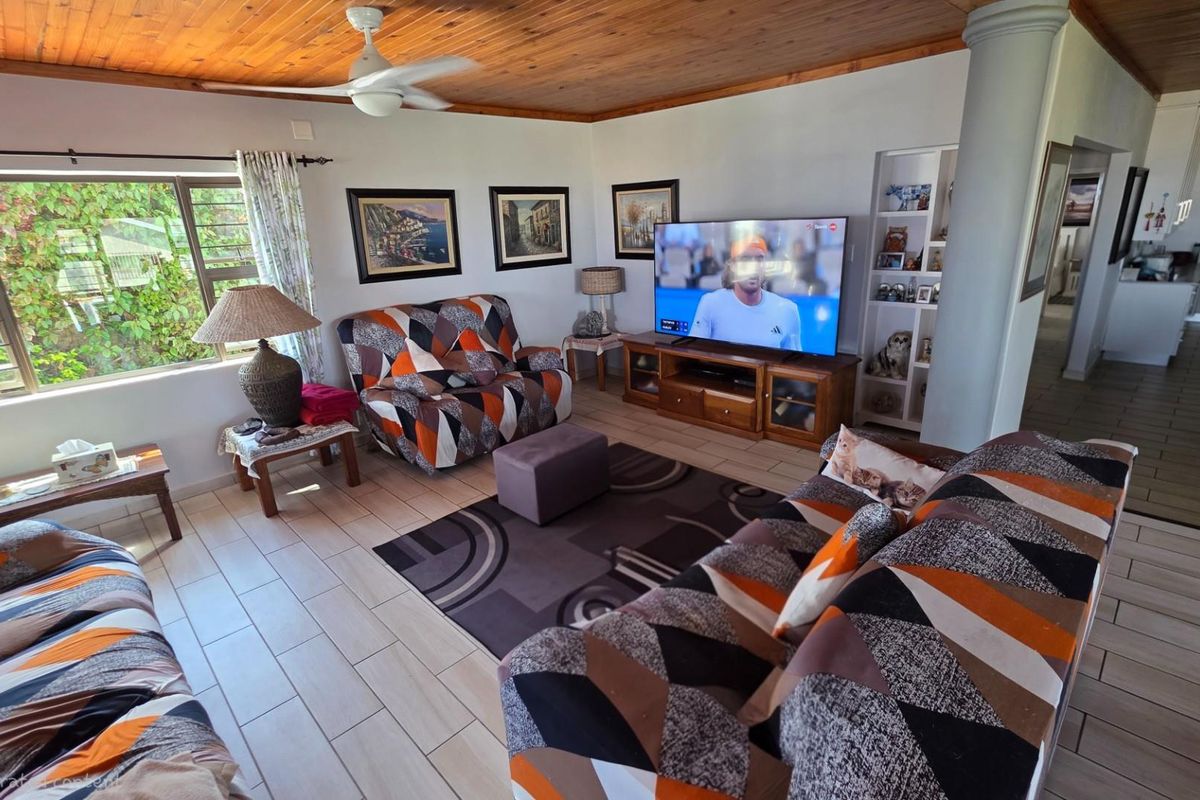 A comfortable living room featuring a timber-clad ceiling, ceiling fan, and tiled flooring, with an open-plan layout leading towards the kitchen area.