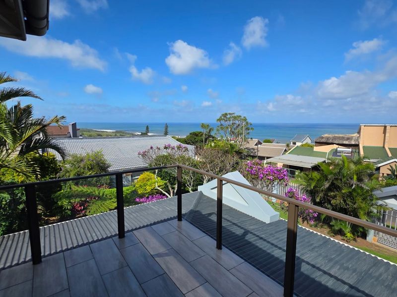 A spacious balcony featuring modern tiled flooring and a sleek glass balustrade, offering expansive elevated views of the surrounding neighborhood and the ocean.