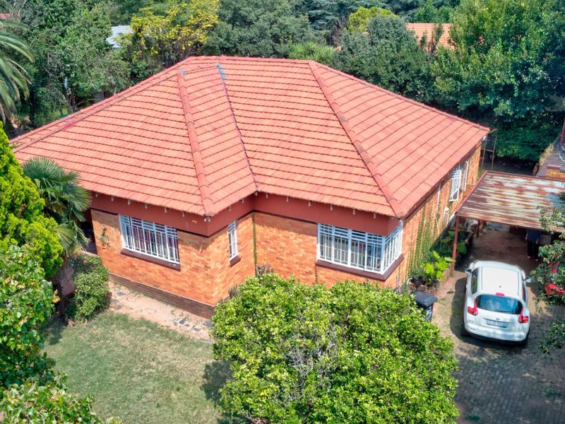Aerial view of a single-story brick house with a tiled roof, featuring a paved driveway and a carport set within a mature garden.