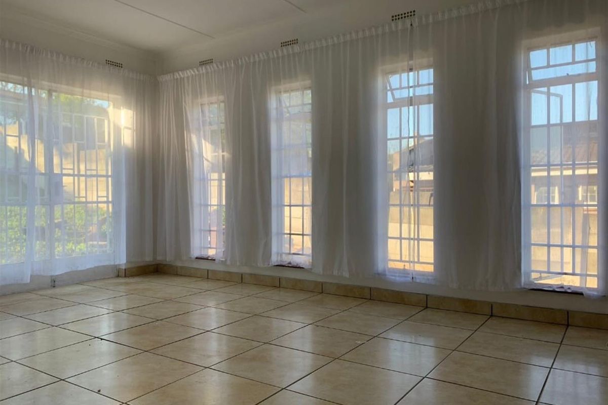 A bright, empty living room featuring large windows with security bars, tiled flooring, and neutral wall finishes.