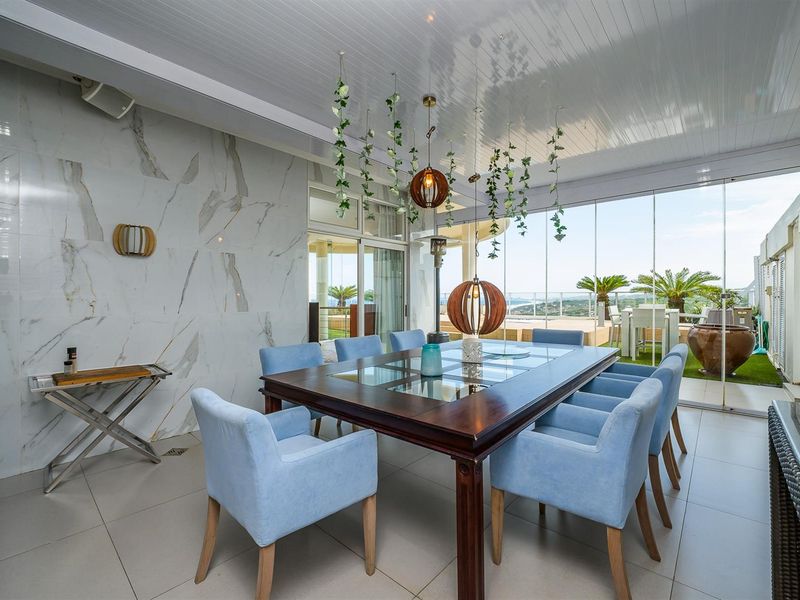 A bright, enclosed patio dining area featuring marble-look wall cladding, tiled flooring, and floor-to-ceiling glass sliding doors offering scenic outdoor views.