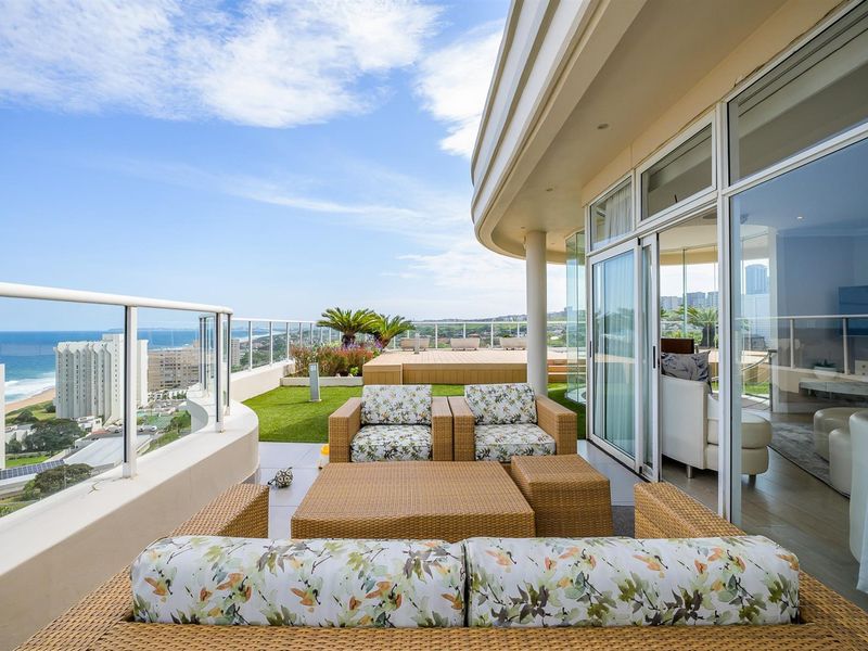 Spacious wrap-around balcony featuring glass balustrades, artificial turf, and panoramic coastal views, seamlessly connected to the interior living space through large sliding doors.
