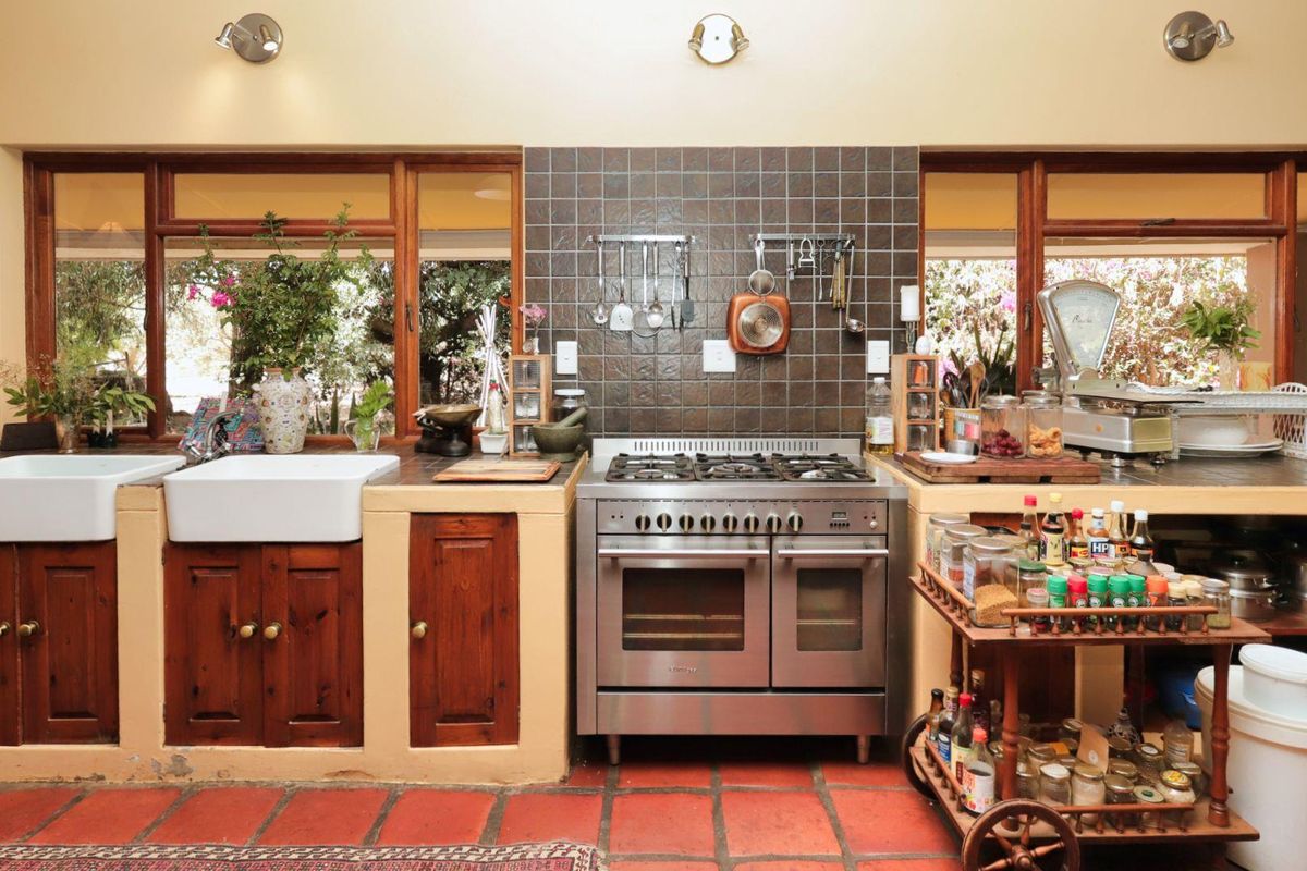 A rustic-style kitchen featuring double farmhouse sinks, a stainless steel gas range, and tiled flooring with large windows overlooking the garden.