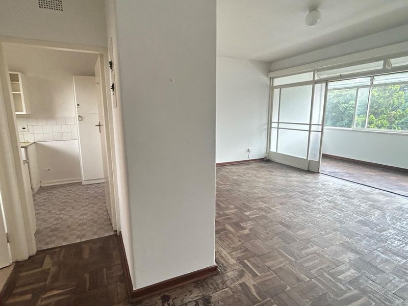 An open-plan living area featuring original parquet flooring, leading to an enclosed balcony with large windows and an adjacent kitchen space.