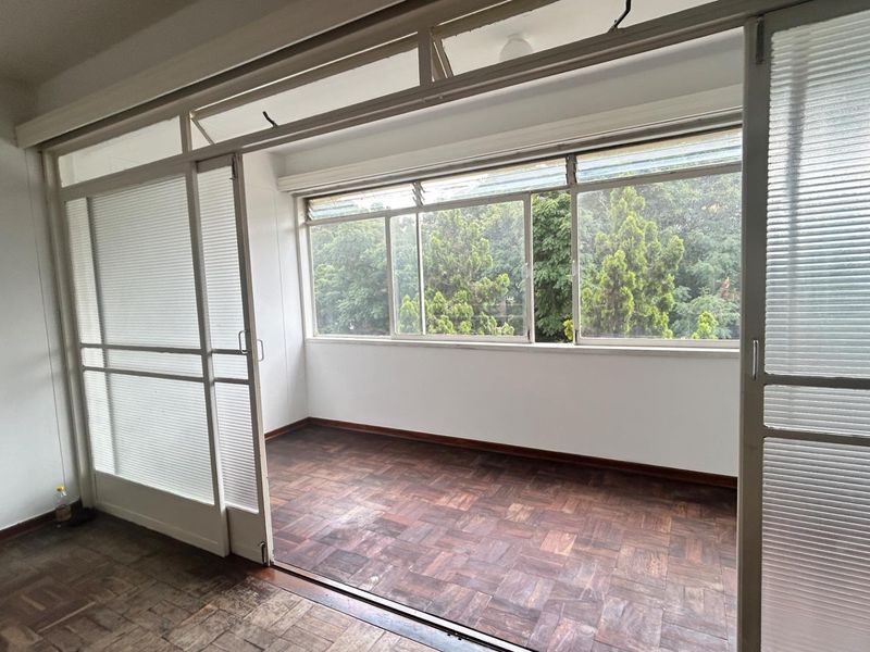 A sunroom or enclosed balcony area featuring parquet flooring, large windows with garden views, and sliding glass partition doors.