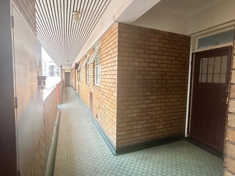 A covered exterior walkway featuring exposed brick walls, tiled flooring, and a slatted ceiling, providing access to commercial units.