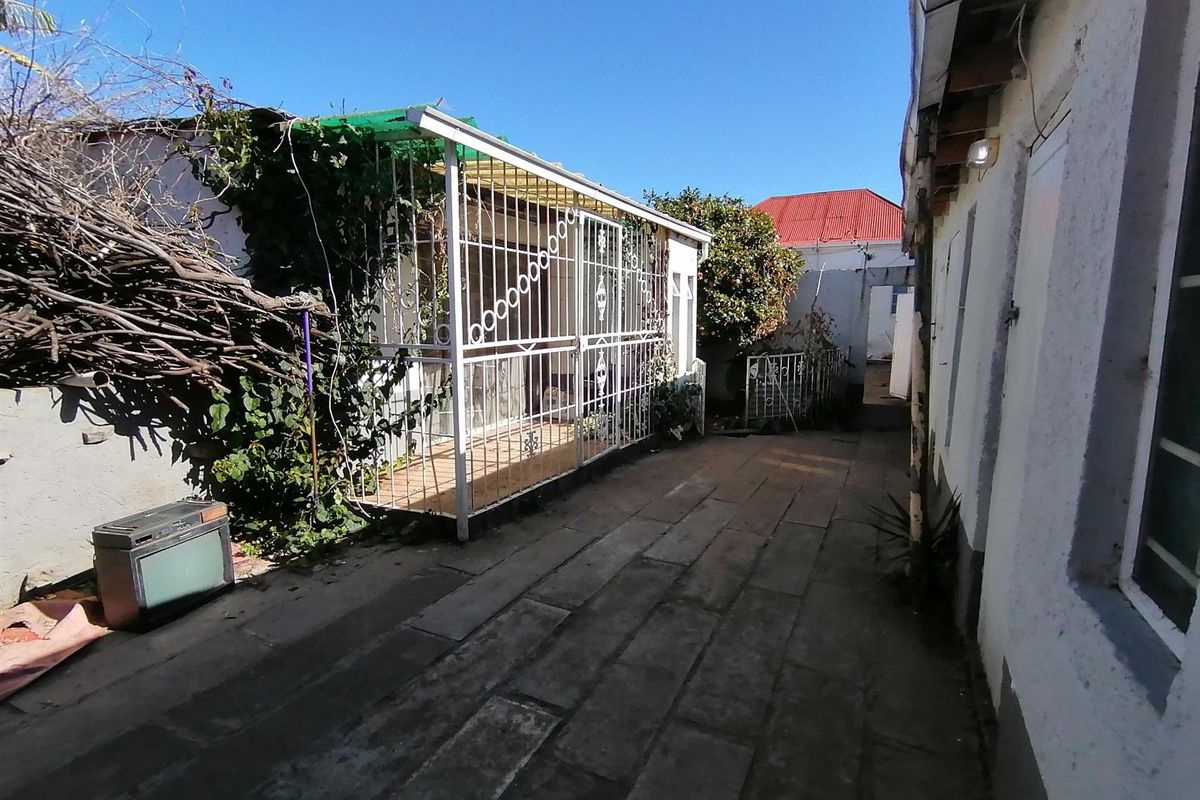 A side exterior area featuring paved ground, a covered patio structure with security bars, and access to the rear of the property.