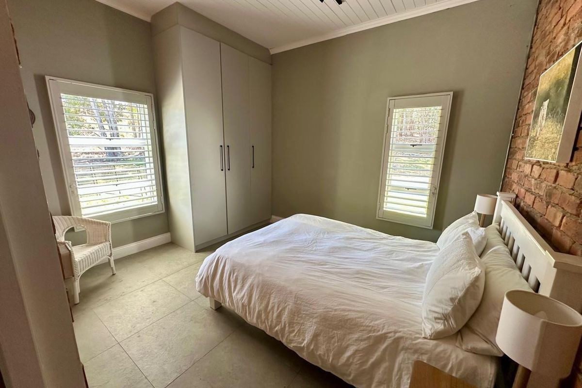 A well-maintained bedroom featuring built-in cupboards, plantation shutters, tiled flooring, and an exposed brick feature wall.