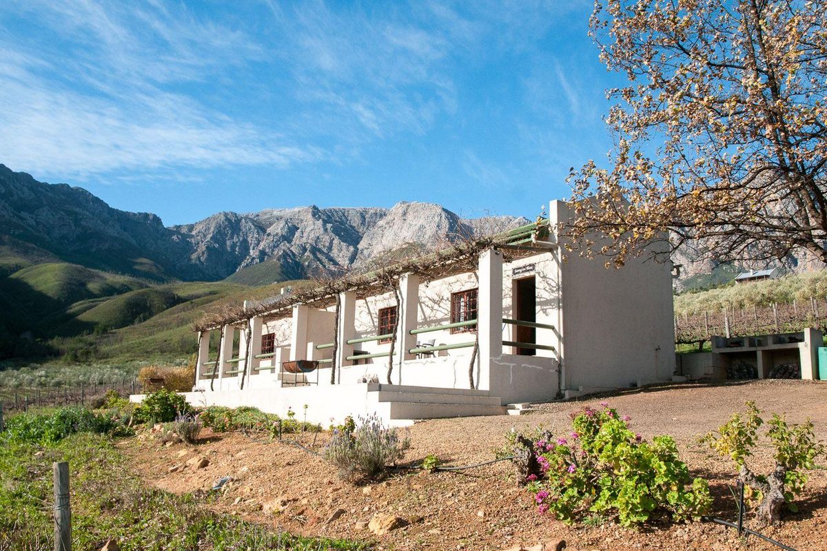 A rustic farm cottage featuring a covered patio with a timber pergola, set against a scenic mountain backdrop with surrounding vineyard views.