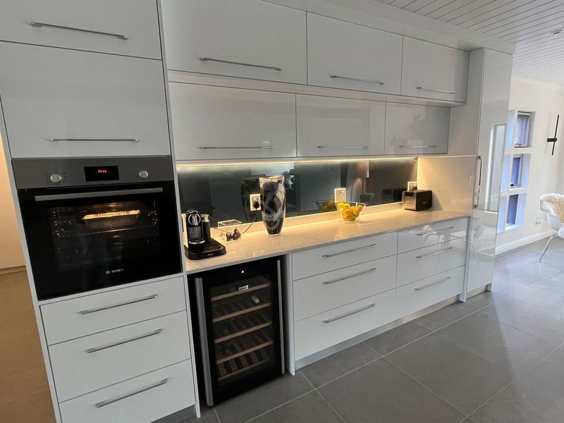 Modern kitchen featuring high-gloss white cabinetry, stone countertops, integrated wine cooler, built-in oven, and sleek glass splashback with under-cabinet lighting.