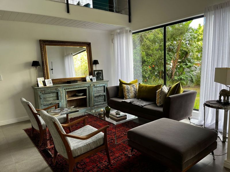A bright, modern living room featuring high ceilings, a mezzanine level, and large floor-to-ceiling windows overlooking a lush garden.