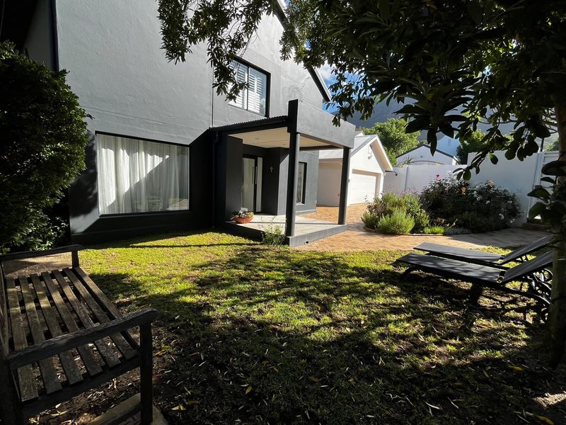 A well-maintained rear exterior view featuring a covered patio, paved area, detached garage, and a landscaped garden with mature trees.