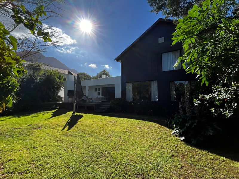Rear view of the property featuring a spacious lawn, timber decking, and an outdoor fireplace, set against a backdrop of mature trees.