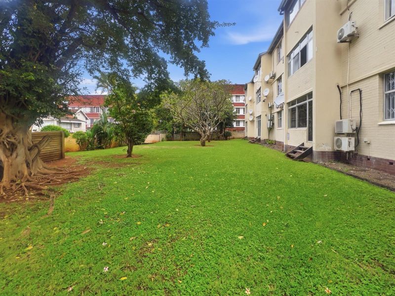 A view of the communal garden area and rear facade of the apartment building, featuring established trees and lawn space.