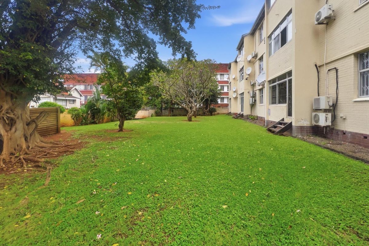 A view of the communal garden area and rear facade of the apartment building, featuring established trees and lawn space.