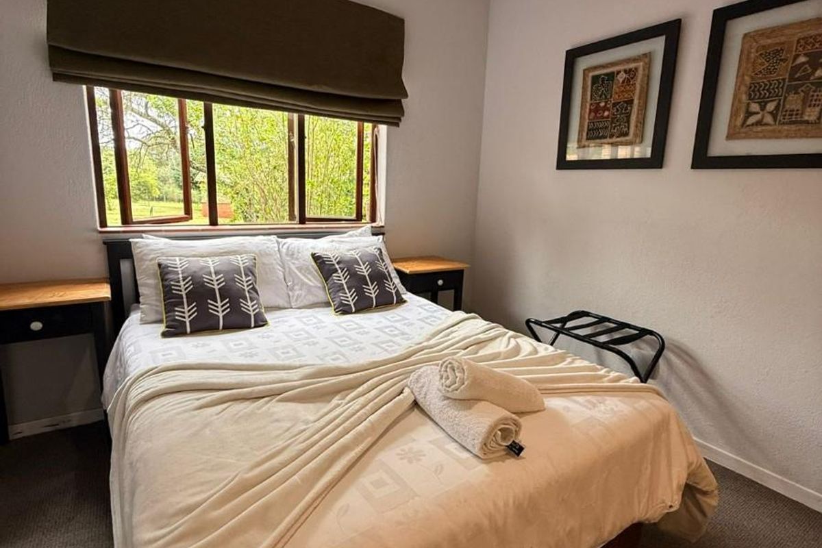 A cozy bedroom featuring a window with a roller blind, a ceiling fan, and neutral carpeting.