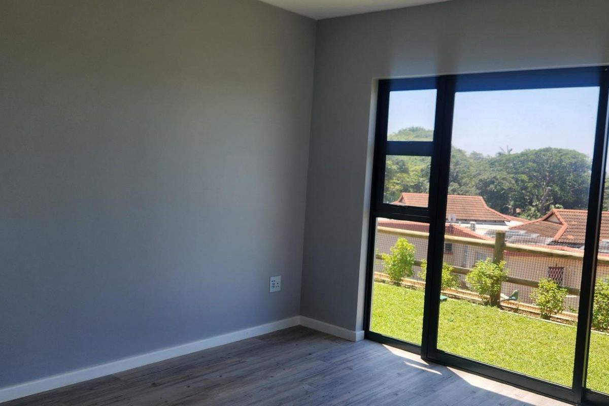 A bright, empty bedroom featuring modern grey walls, wood-look flooring, and large sliding glass doors that open to a green outdoor view.